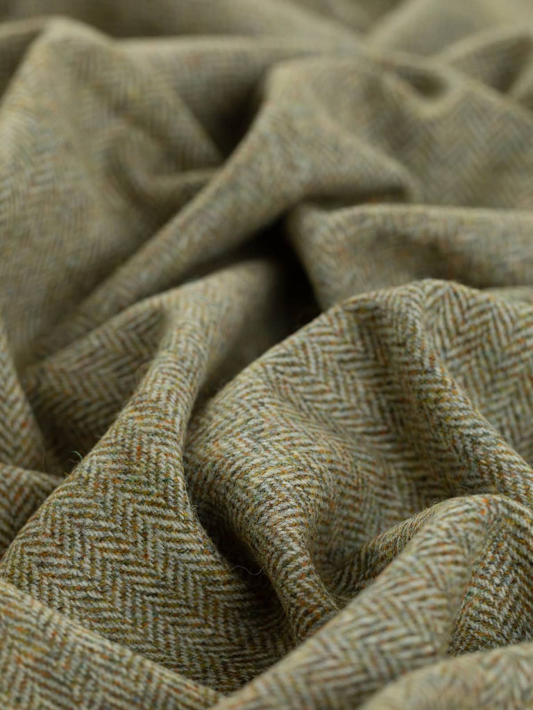 A close-up view of Marvellous Moss fabric twisted to showcase the rich herringbone pattern. The melange of golden olive, rust, and blue-green weft yarns creates a moss-like appearance, contrasted by light grey marled warp yarns. The chevron detail is highlighted, illustrating the fabric’s natural, earthy tones.