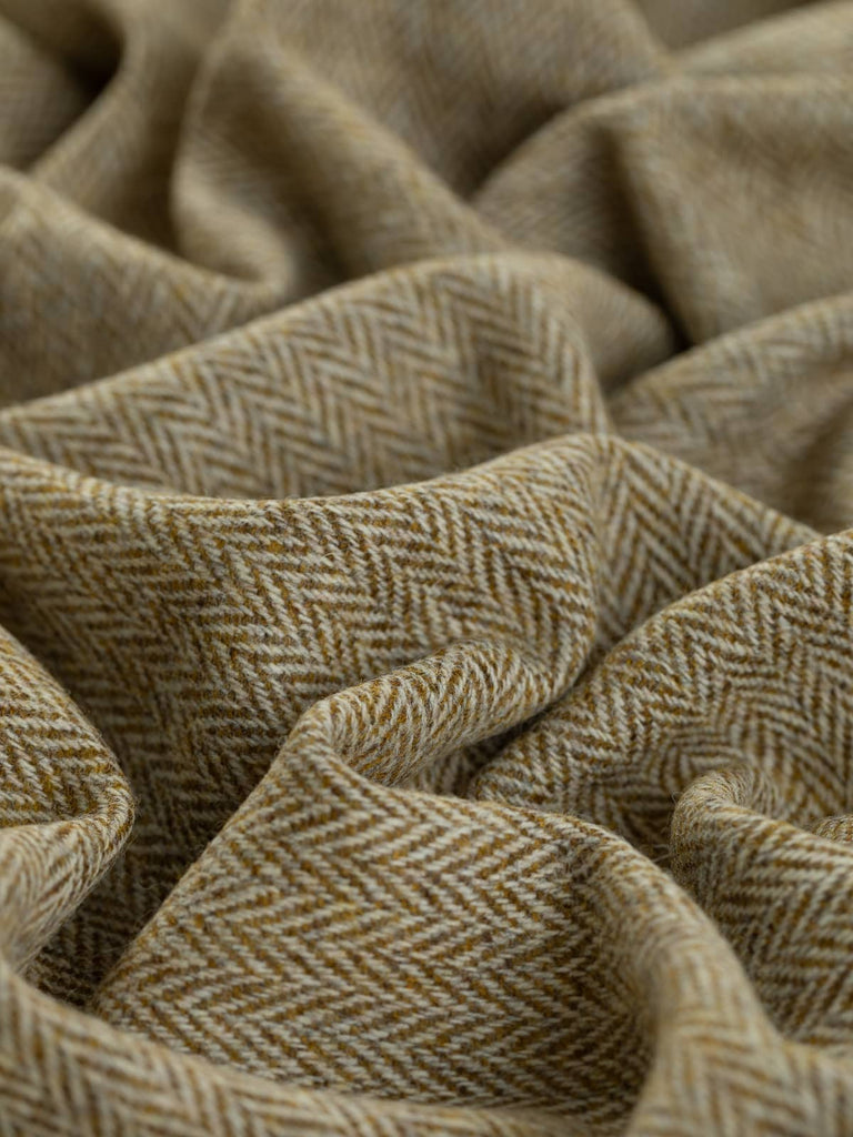  A close-up shot of the Yorkshire Tweed Herringbone - Turning Seasons, twisted to emphasize the intricate herringbone pattern. The fabric’s rich, golden olive green weft intertwines with natural white warp yarns, evoking the warm tones of late summer leaves transitioning into autumn. The subtle chevron design highlights the exquisite craftsmanship of this luxurious wool tweed.