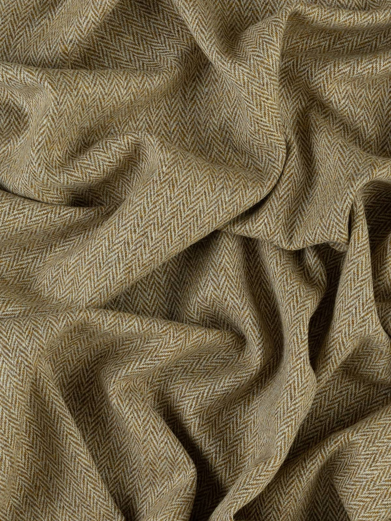 An aerial view of the Turning Seasons herringbone tweed, gracefully twisted to display its sun-kissed golden olive tones and soft chevron pattern. The image captures the fabric’s luxurious wool texture and stable drape, evoking the warmth and elegance of autumn’s first days. A versatile fabric for both fashion and interior design, with a nostalgic, nature-inspired color palette.