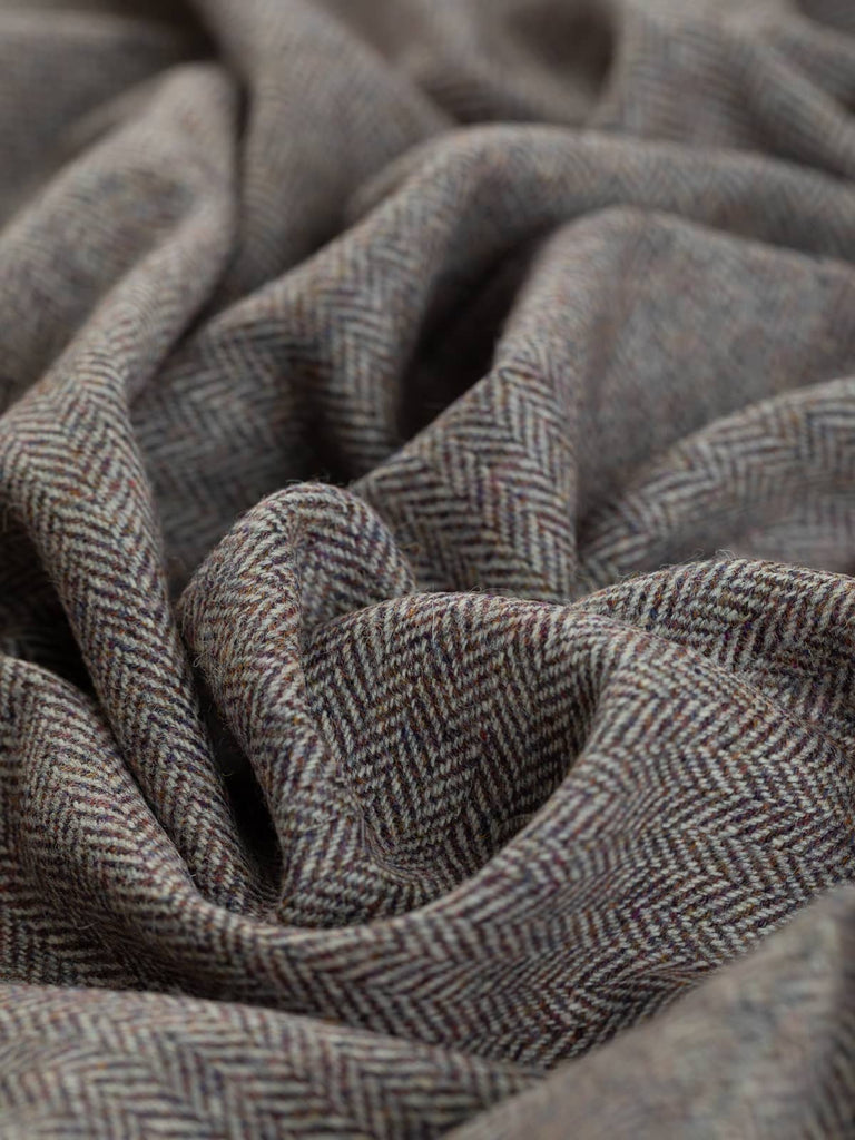 A close-up view of the Heather Momento fabric, twisted to showcase the intricate herringbone pattern. The blend of heather purple, rust, and autumn gold hues is interwoven with marled natural grey, creating a rich, textured appearance reminiscent of the North York Moors. The fine weave and classic chevrons highlight the fabric's natural elegance.