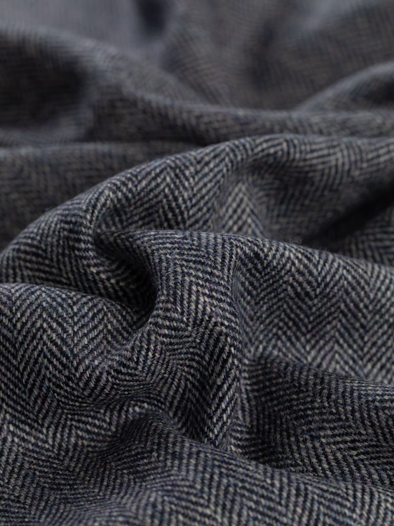 A detailed close-up of the Blackthorn Blue Herringbone Tweed, capturing the intricate combination of soft heathered lilac and natural grey yarns. The characteristic chevron pattern is sharply defined, reflecting the subtle variations in tone and texture that evoke the timeless elegance of classic tweed, inspired by the deep purplish-blue of blackthorn berries.