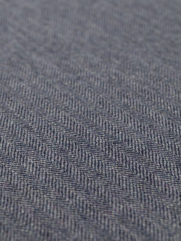 A flat angled view of the Eco Herringbone Yorkshire Tweed in Blackthorn Blue, showcasing its subtle chevron pattern. The fabric reveals a beautiful interplay of dark navy and lilac undertones, combined with natural sheep beige and marled grey yarns, creating a misty lilac-grey haze. The intricate herringbone weave is visible, adding texture and depth, perfect for classic and contemporary tailored pieces.