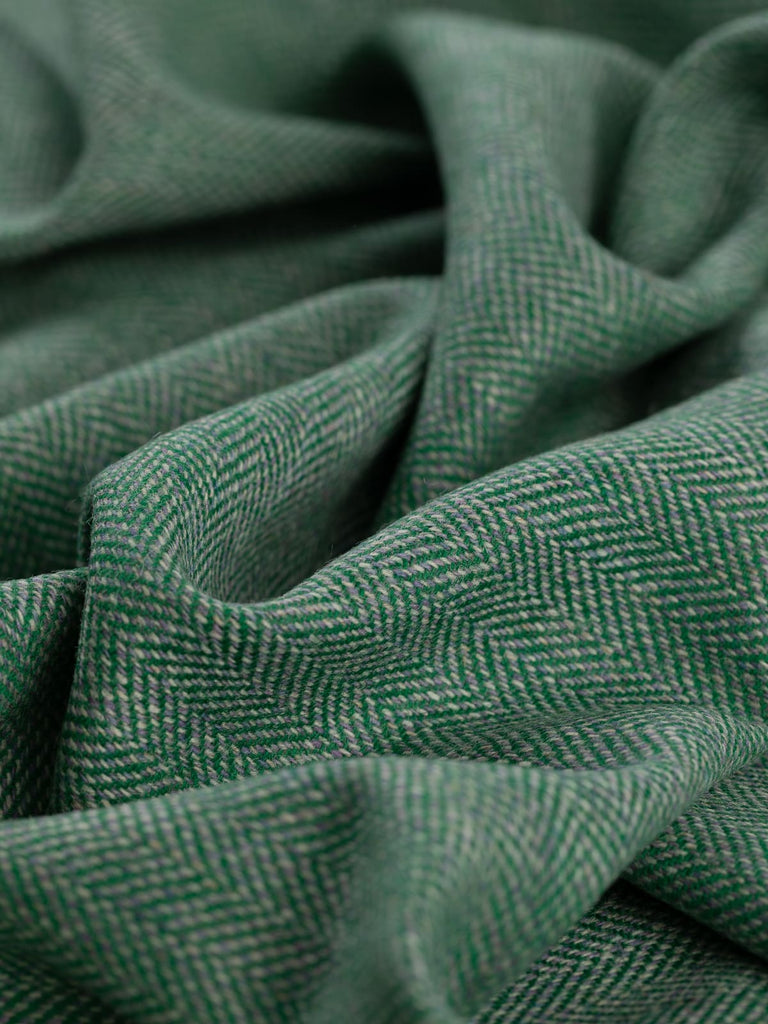 Angled close-up of the twisted Eco Herringbone Yorkshire Tweed in Verdant Valley, emphasizing the lively emerald green chevrons and the soft lilac highlights. The texture and intricate pattern of the fabric are showcased.