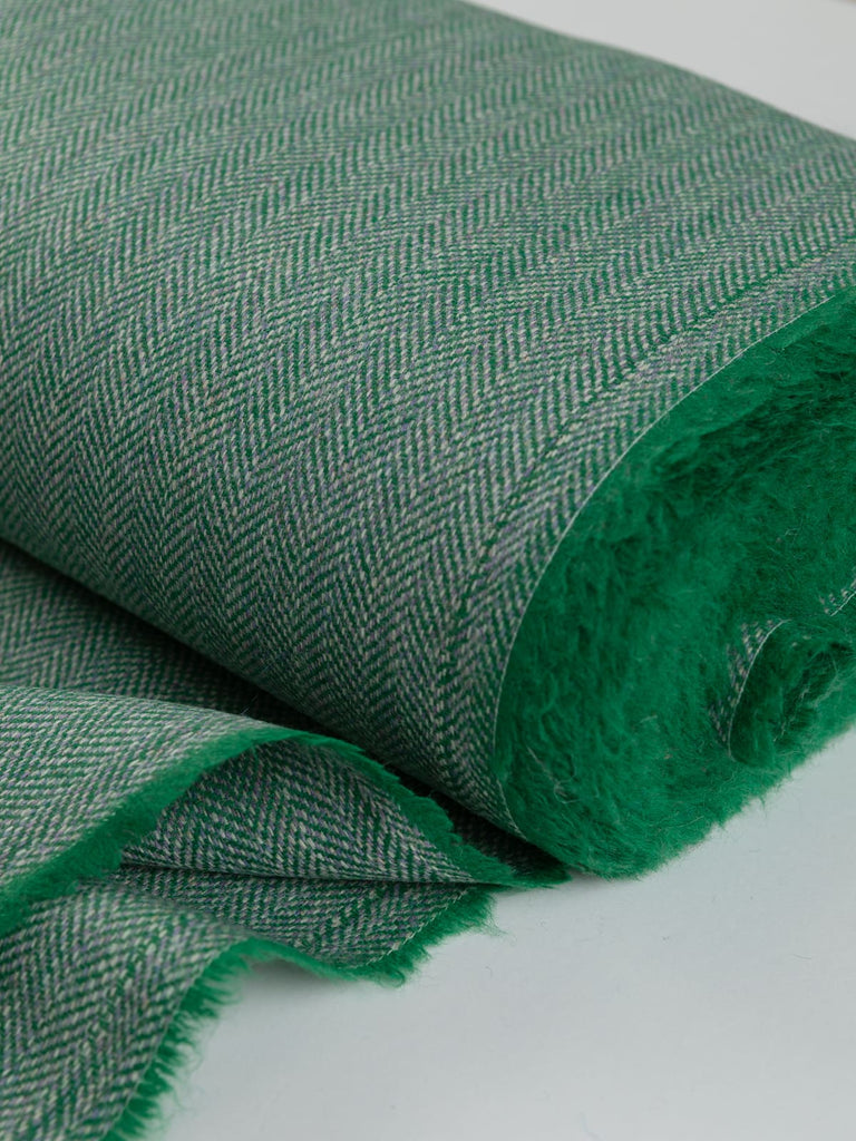 Close-up of the bolt end of Eco Herringbone Yorkshire Tweed in Verdant Valley, showing the selvedge and the vibrant glaucous green fabric with lilac undertones. The detailed herringbone pattern and rich texture are clearly visible.