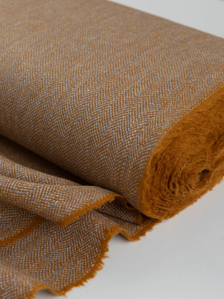 Close-up of the bolt end of Eco Herringbone Yorkshire Tweed in Barley Bounty, highlighting the golden ochre fabric with lilac undertones. The selvedge and distinctive herringbone pattern are clearly visible, showcasing the texture and warm hue