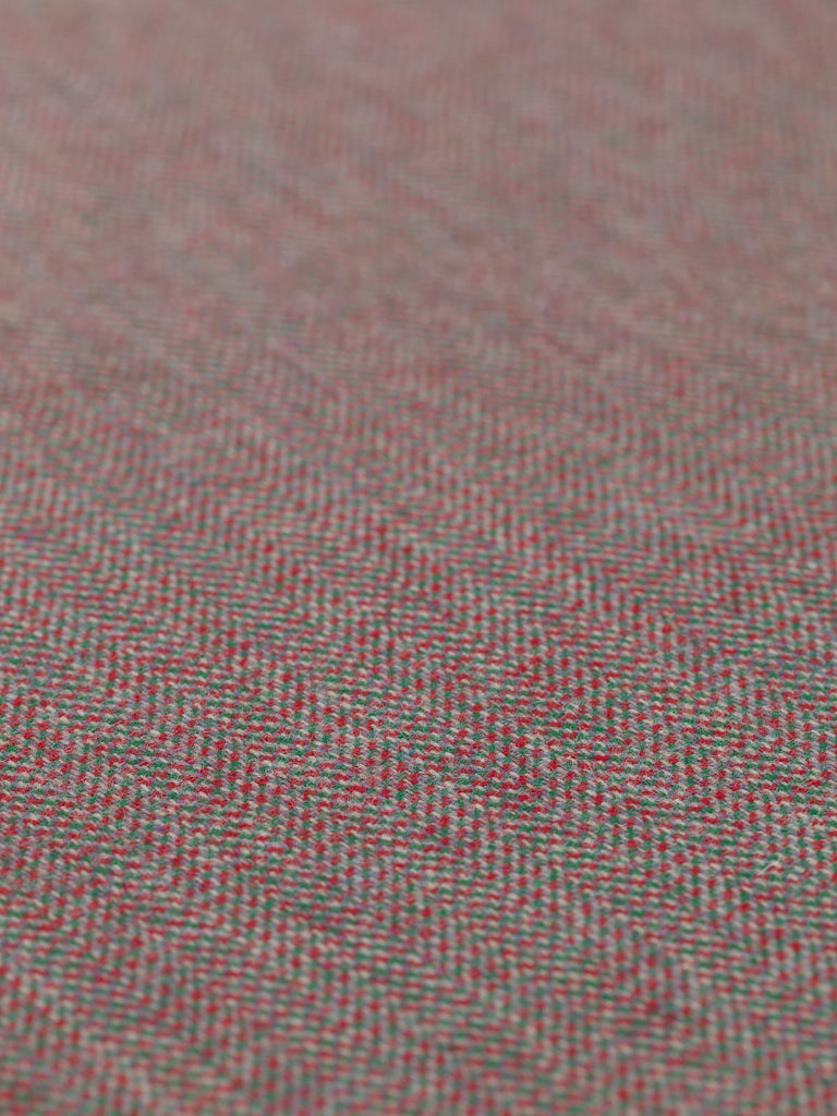 Angled detailed close-up of Eco Herringbone Yorkshire Tweed in Festive Folly, focusing on the flat weave and alternating red and green with lilac undertones. The herringbone pattern and festive color scheme are clearly visible.