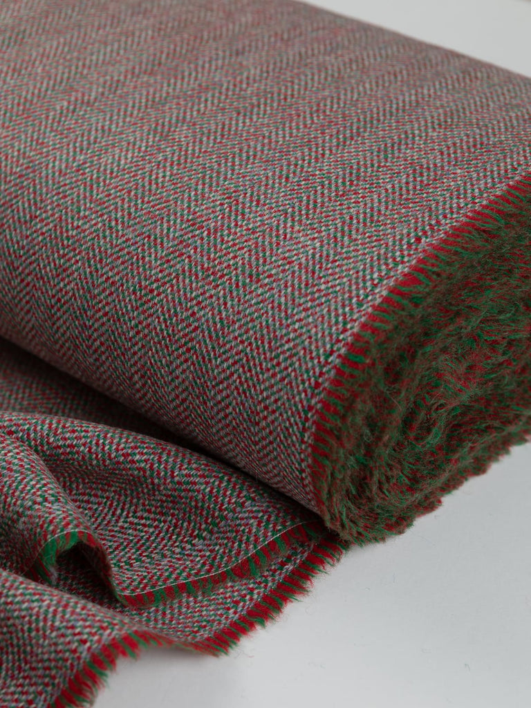 Close-up of the bolt end of Eco Herringbone Yorkshire Tweed in Festive Folly, featuring festive red and green with lilac undertones. The selvedge and herringbone pattern are highlighted, capturing the vibrant and cheerful fabric.