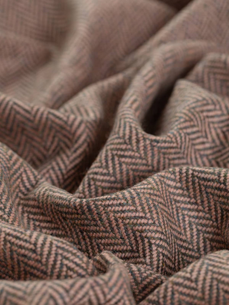 A close-up view of the Nostalgic Blush fabric, twisted to showcase the medium/large herringbone pattern. The blend of smoke grey melange yarns, with hints of blue-grey, loden green, and subtle brown-heather tones, is beautifully contrasted by a pale vintage powder pink weft. The soft, luxurious texture reflects the superior quality of lambswool and evokes a nostalgic, vintage charm.