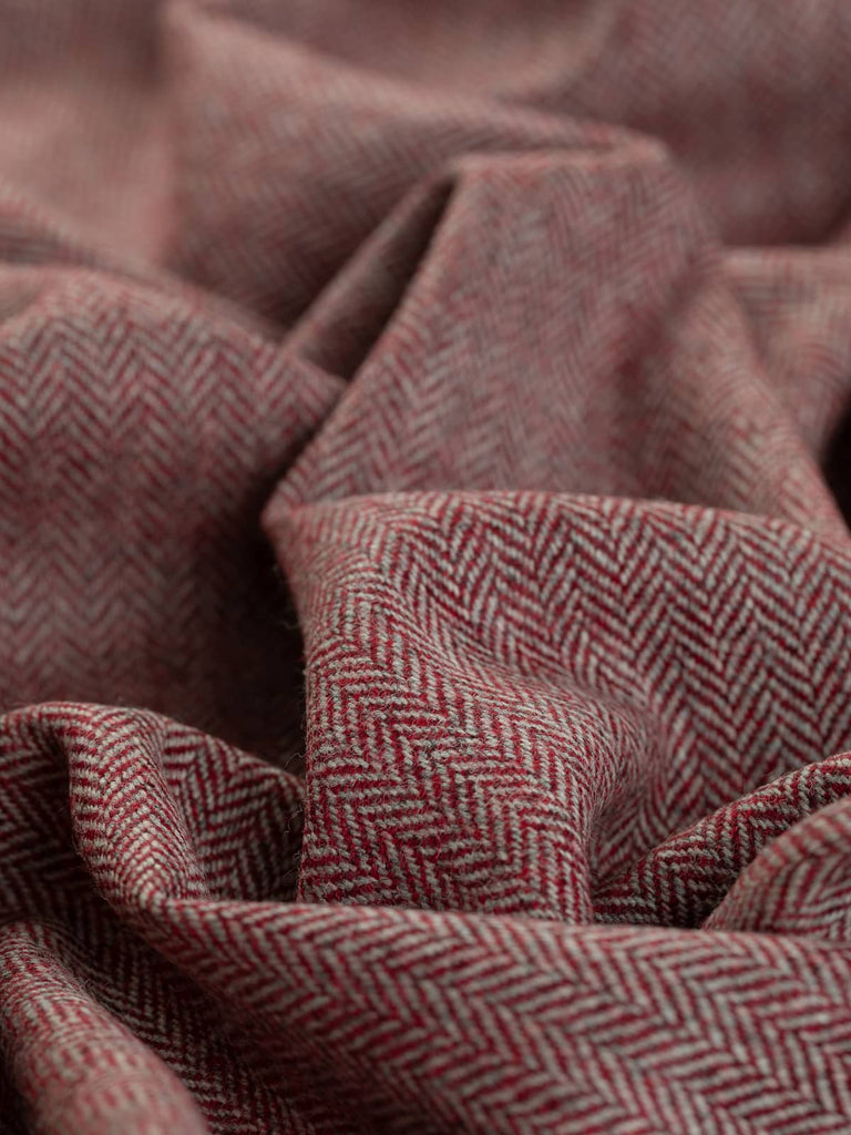 A close-up of the Lifeblood fabric, twisted to highlight the classic fine herringbone weave. The rich melange of red yarns, reminiscent of blood-red winterberries, contrasts beautifully with marled natural grey warp yarns. The intricate chevron pattern and vibrant red tones evoke a sense of tradition and warmth, making the fabric perfect for both clothing and interiors.