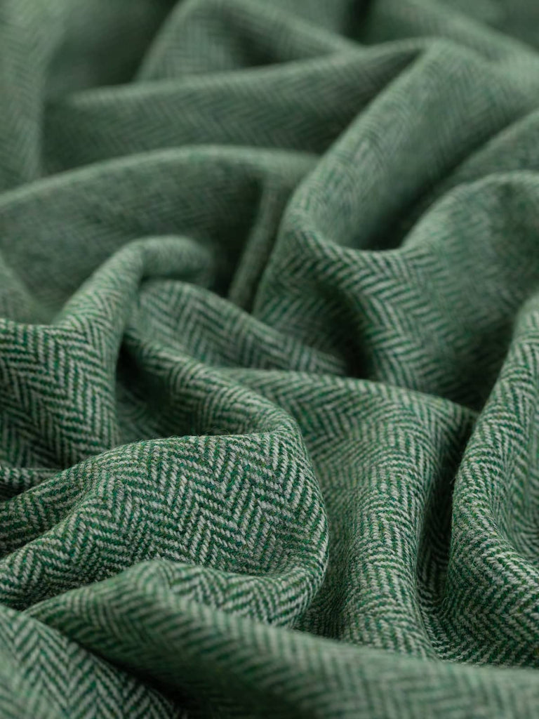 A close-up of the Green Pastures fabric, twisted to showcase its classic herringbone pattern. The deep, chlorophyll green weft yarns contrast subtly with the soft marl grey warp yarns, evoking the lush look of summer grass. The intricate chevron detailing adds depth and texture, highlighting the fabric's natural and vibrant green tones.