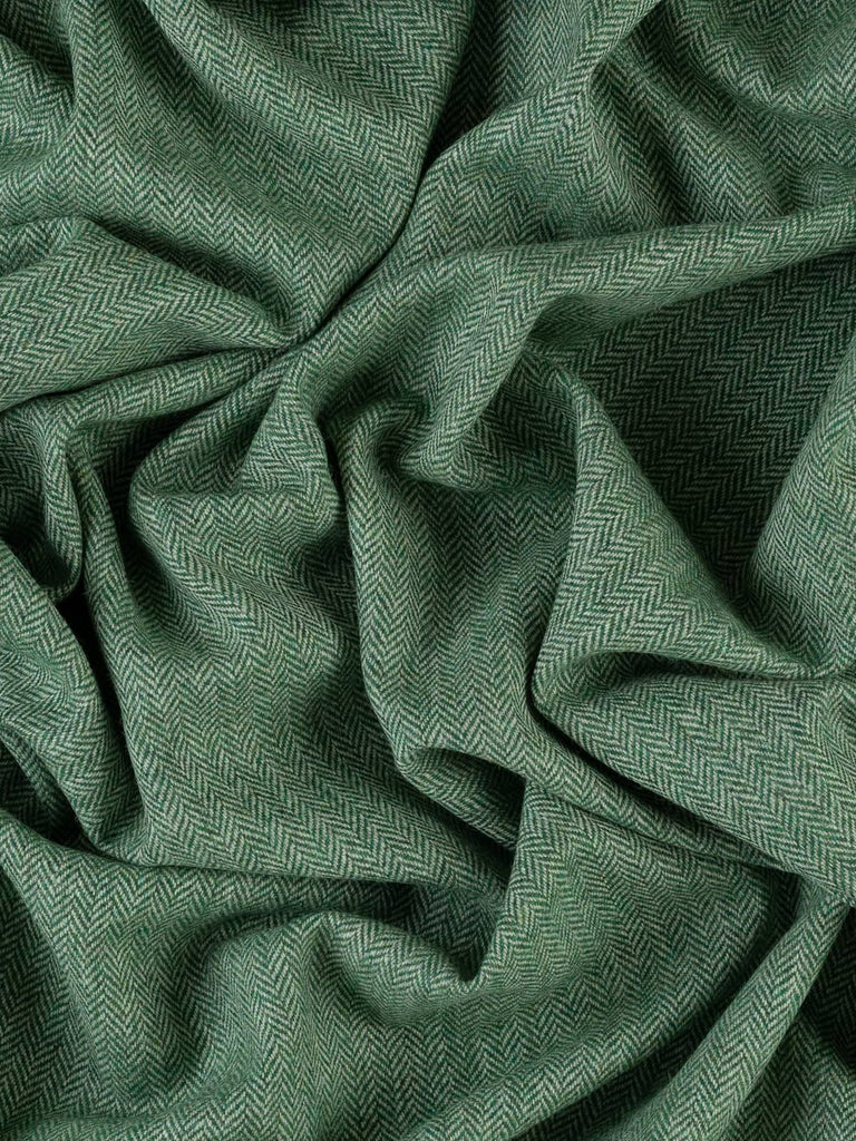 An aerial view of the Green Pastures fabric, twisted to enhance the chevron pattern and the dynamic blend of green and grey tones. The deep, lush green hues are reminiscent of grassy summer fields, with subtle grey contrasts that define the herringbone design. The fabric’s soft texture and versatile medium weight make it ideal for both garments and home furnishings.