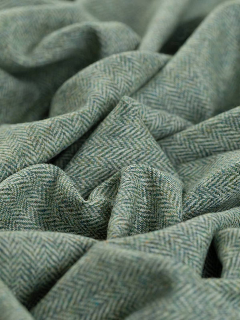 A close-up view of Moonlit Lichen fabric twisted to highlight the intricate herringbone pattern. The multi-tonal blue-green melange weft yarns give the fabric a frosted, lichen-like appearance, contrasted with subtle grey tones that evoke moonlit dew. The detailed chevron pattern is clearly visible, showcasing the fabric’s unique texture.