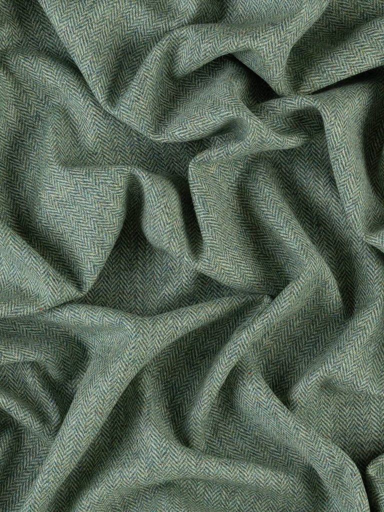 An aerial view of Moonlit Lichen fabric, twisted to display the chevron pattern and the blend of frosted blue-green and grey hues. The fabric’s stylish, fresh appearance and its texture, reminiscent of lichen-covered branches, are highlighted from this angle. The medium weight and classic tweedy feel are apparent.