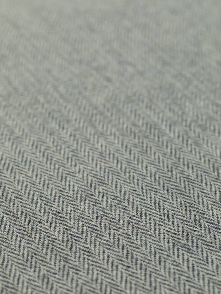 A flat, angled view of Tempest fabric, capturing the interplay between the cool storm grey and dark navy blue yarns. The herringbone pattern is prominently displayed, illustrating the fabric’s classic tweedy texture and medium weight. The contrast between the hues evokes the feeling of a turbulent ocean storm.