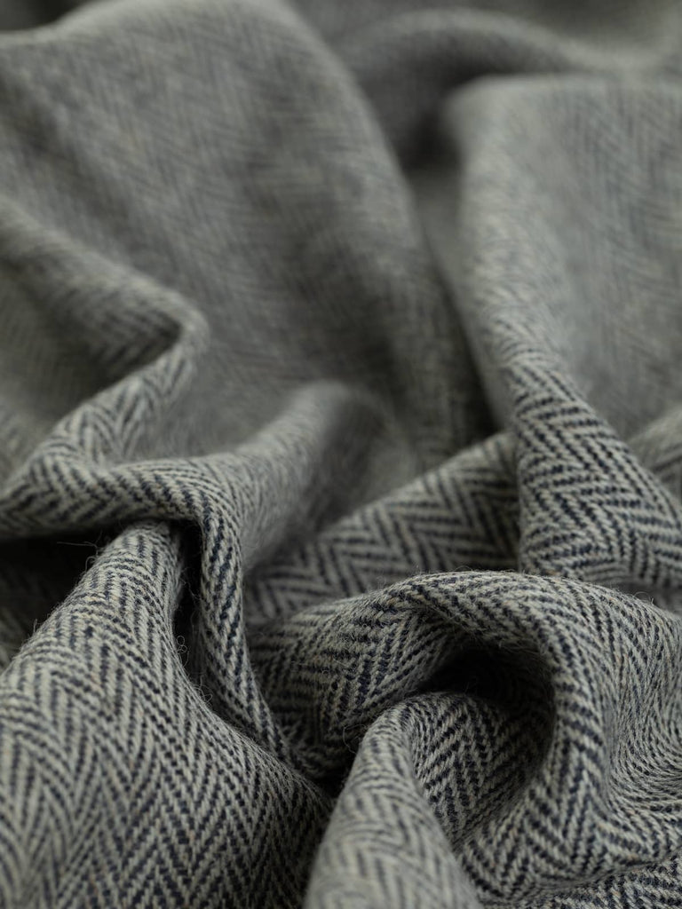 A close-up view of Tempest fabric twisted to showcase the fine herringbone pattern. The fabric features a storm grey warp melange yarn base and deep navy blue weft yarns, creating a dramatic contrast that highlights the tempestuous, lightning-like chevrons. The texture and pattern are emphasized from this angle.