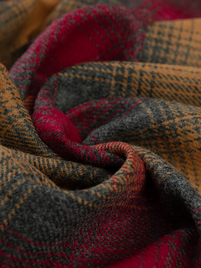 Angled twist shot of Fireside Glen Check Yorkshire Tweed, showing grey-green melange base with glowing amber and ember-red twill lines inspired by a warm hearth.
