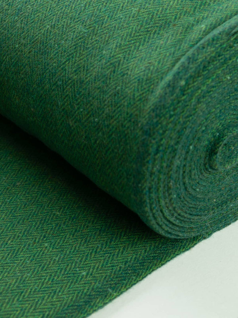 Angled flat view of luxurious Irish Donegal Tweed in a deep resinous pine green, showcasing an intricate herringbone pattern enhanced by fresh grass-green melange yarns and rustic flecks of turquoise, beige, and rust.