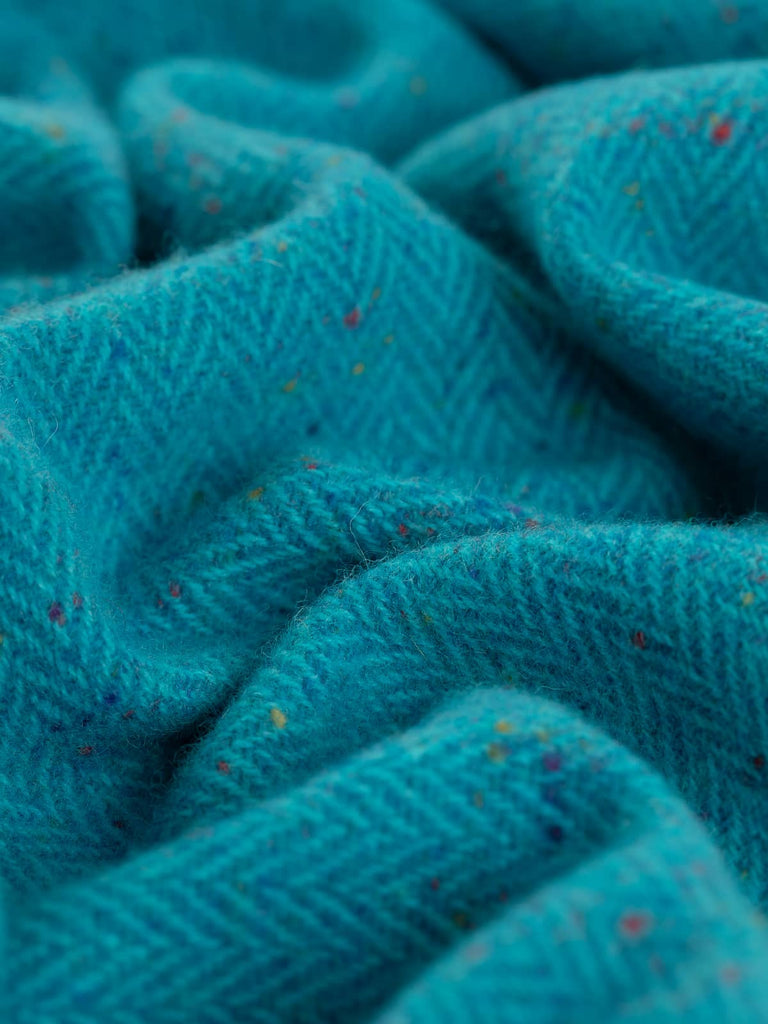 The close-up shot of the Electric Turquoise fabric reveals a lively interplay of vibrant turquoise hues and rainbow Donegal flecks, showcasing the dynamic texture and energy of this first-rate Irish Donegal Tweed Herringbone.