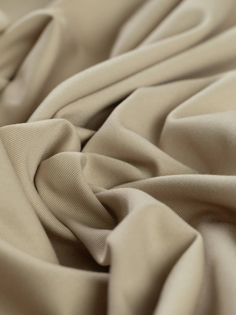 Angled twist shot of Sandstorm fabric, showing soft folds that emphasize its fluid drape and pale chino beige color. The fine twill weave and slightly lustrous finish give the fabric a smart, understated character with a natural, earthy feel.