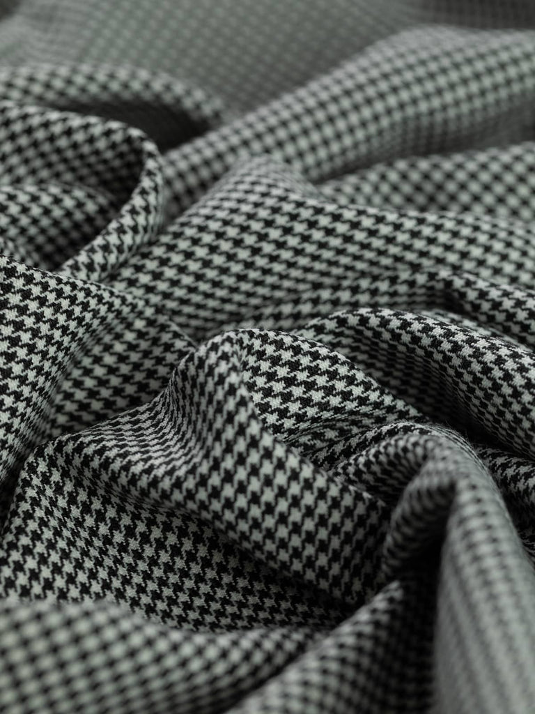 Angled twist shot of the fabric showing the small-scale black and white houndstooth pattern, with soft folds enhancing the lightweight drape and tailored structure. The fine weave highlights the fabric’s high-quality texture and smart, vintage-inspired aesthetic
