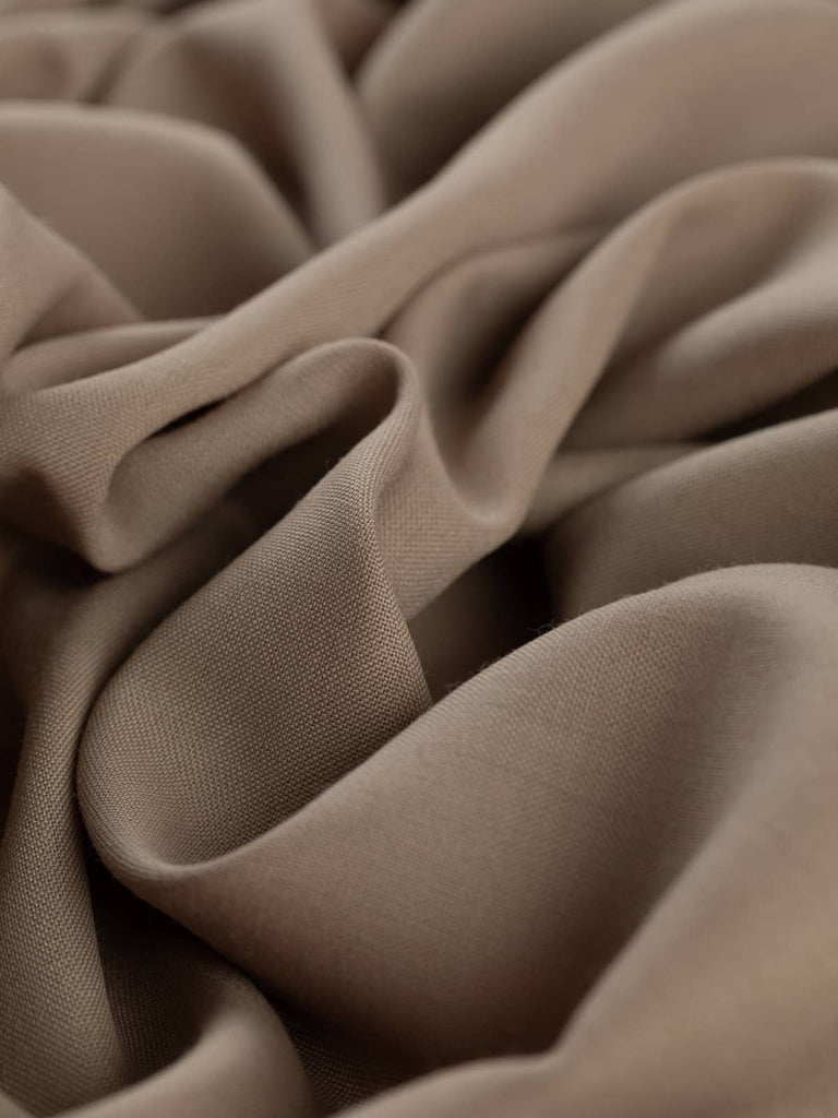 Twisted, angled shot of Hazel Wood fabric, highlighting the warm beige hue and fluid drape. The fine flat weave texture appears smooth and refined, emphasizing the fabric’s soft, silky feel and its suitability for tailored or chic minimalist garments.