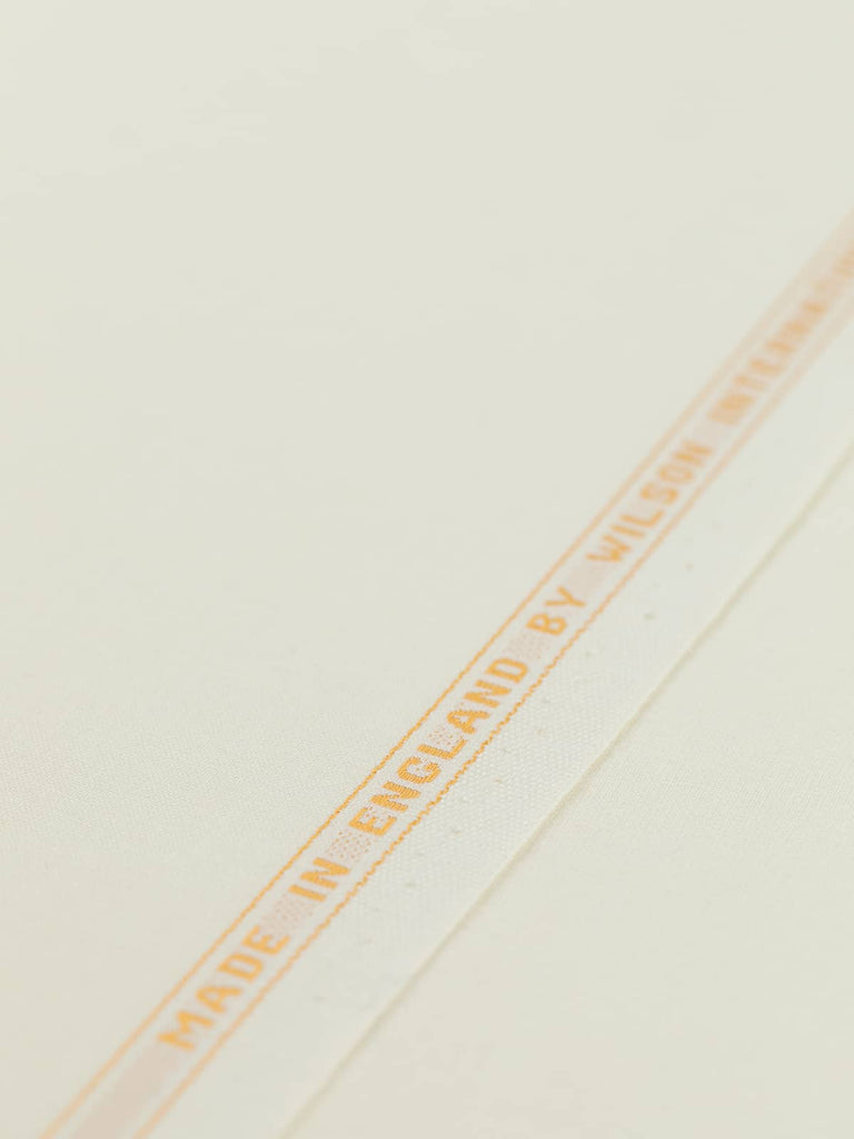 Flat angled view of Classic Ecru wool-blend worsted fabric, highlighting its pale cream-white tone, golden selvedge, and crisp yet elegant drape
