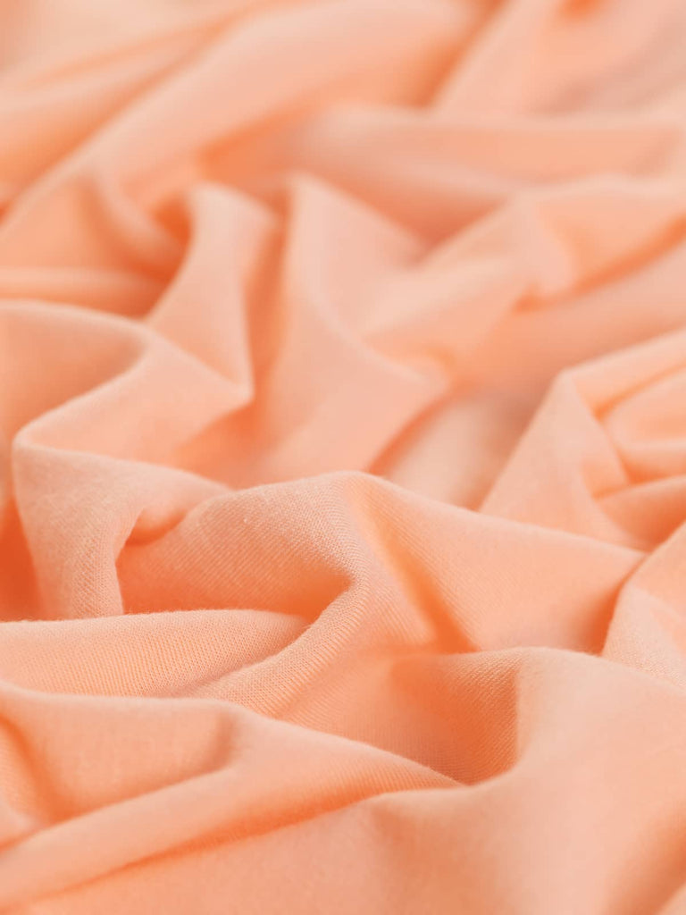 A close-up of the Cosy Soft Single Jersey fabric in Fuzzy Peach, highlighting its pastel peachy pink-orange tone and almost brushed texture. The twisted view reveals the fabric's cosy warmth and excellent retentive stretch, making it perfect for soft and comfortable garments.