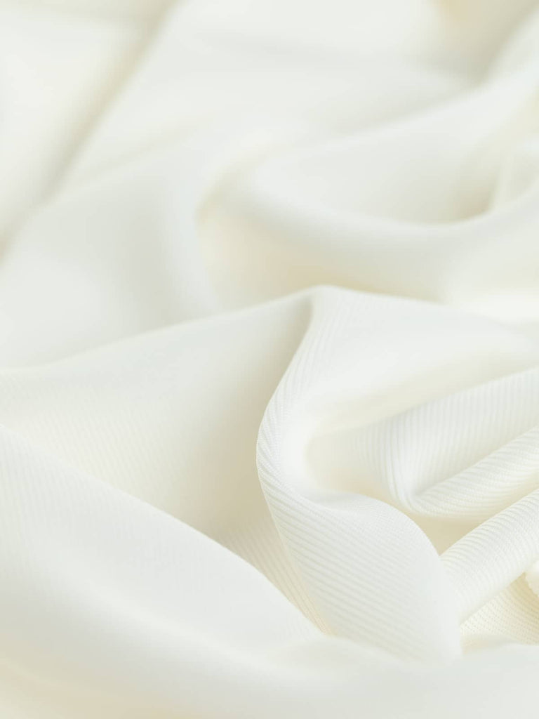 A close-up of the Slinky Stretch Rib fabric in Milk White, highlighting its fine knit and soft, slightly grainy texture. The twisted view reveals the fabric's practical stretch and smooth drape, perfect for creating versatile and elegant garments.