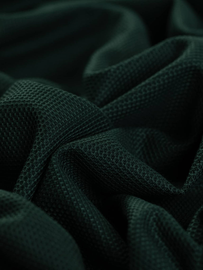 A close-up of the Pique Interlock Jersey in Green Peaks, showcasing its finely knitted diamond honeycomb texture and deep evergreen hue. The twisted fabric highlights its medium weight and natural stretch, perfect for stylish and breathable garments.