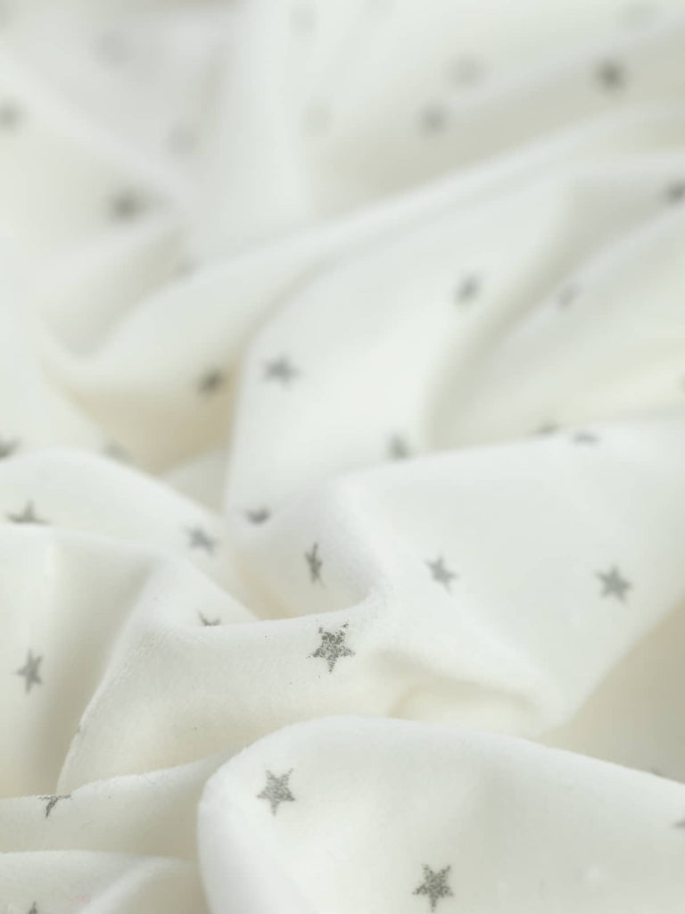 Snow & Stars Cotton Velvet twisted to highlight its soft, plush texture and elegant drape. The pure white cotton base is adorned with silver-grey printed stars with a subtle metallic sparkle, making it ideal for festive and winter-themed garments.