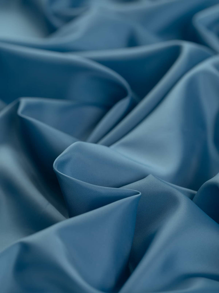 Scandi Blue woven lining fabric, 140cm wide, in a superfine solid flat weave with a cool mid-blue tone and grey undertones. Made from 100% polyester, this very light fabric is soft, silky, and smooth with a semi-shiny, satin-like finish, perfect for lining jackets, skirts, and stylish fashion pieces.