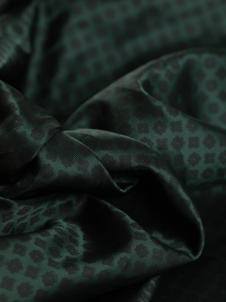 A close-up view of the Viscose Twill Lining - Quatrefoil and Star, showcasing the intricate geometric pattern of quatrefoil and star motifs. The twisted fabric highlights the soft, silky, and lustrous nature of the 100% viscose material. The dark spruce green color with matte black print creates a sophisticated contrast, perfect for high-quality garment linings. 