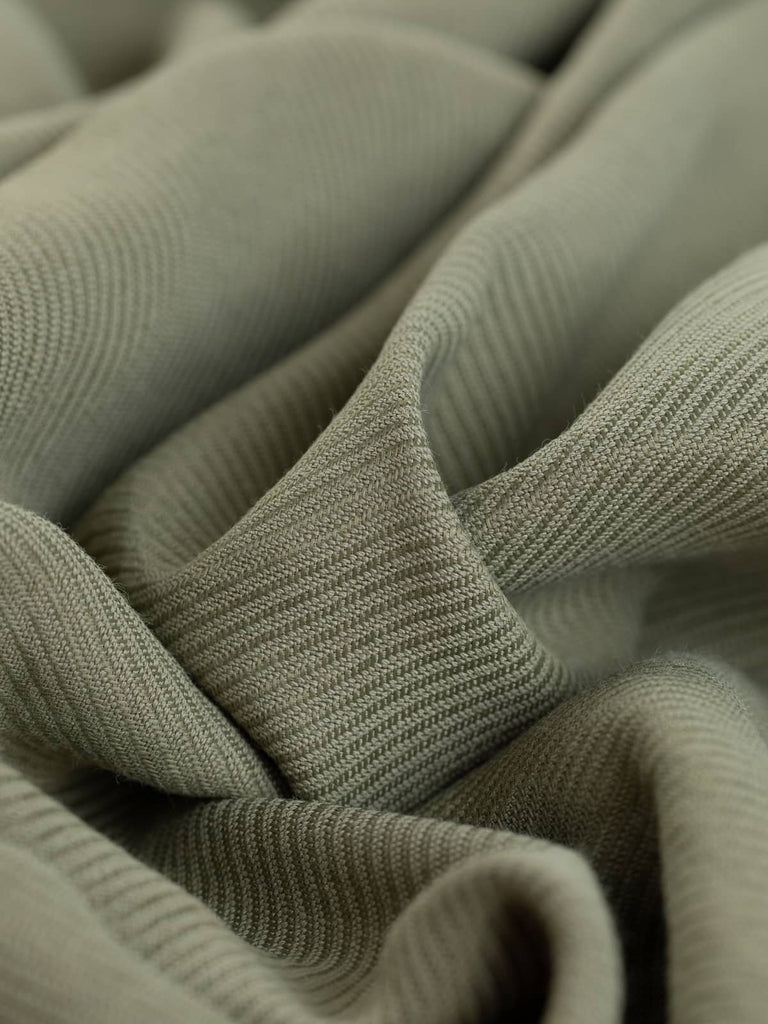 A close-up of the Chunky Viscose Twill - Herb Sachet, showcasing the intricate texture of the chunky twill weave. The twisted fabric highlights the medium weight and the slightly raised ridges, emphasizing the natural green/beige hue reminiscent of dried herbs. This view demonstrates the fabric's soft and malleable flowing drape quality, attributed to the viscose content, and its stable, sturdy hand due to the cotton composition.