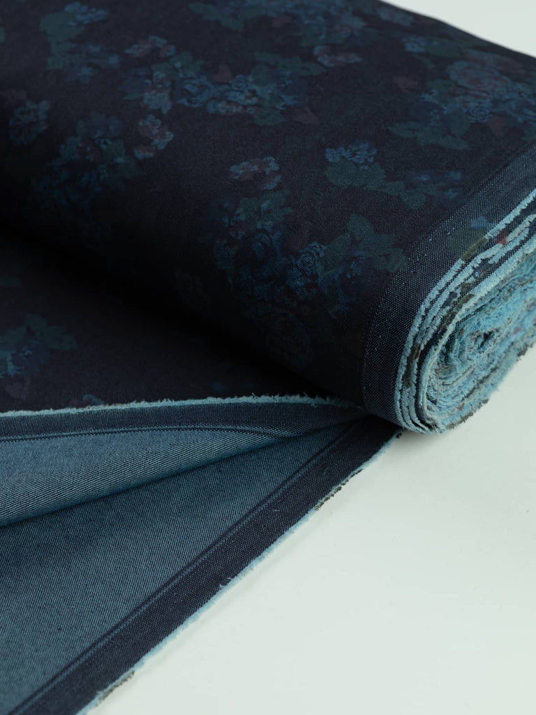 A neat roll of Indigo Rose denim, showcasing its dark indigo base and delicate vintage rose print in shades of teal and mauve. The medium-weight denim has an absorbent cotton hand, and a touch of elastane provides added stretch, perfect for fitted styles like jeans, skirts, and dresses.