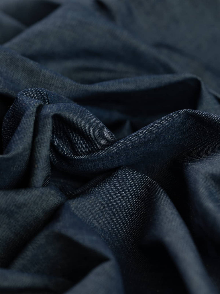  A shot of Dark Indigo chambray denim twisted to show its fine diagonals of dark indigo navy yarns on the warp and soft ecru cotton weft. The lightweight denim has a stable yet soft hand, perfect for creating delicate gathers, tucks, and tiered dress styles.