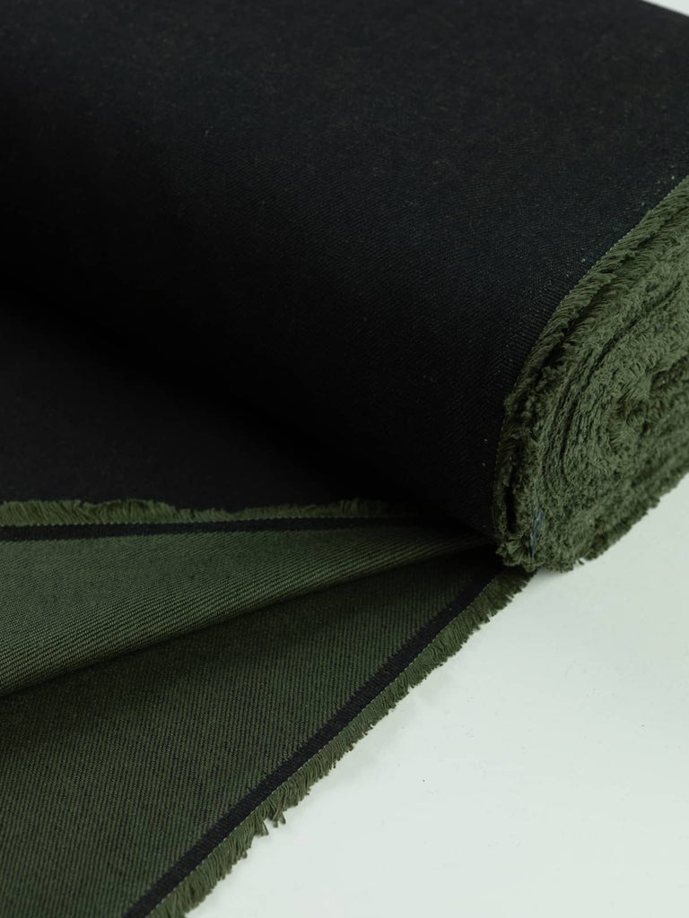 A neatly rolled piece of Khaki Shot Denim fabric, displaying the contrast between its dark green-black face and softer khaki-toned reverse. The sturdy, absorbent cotton weave and fine twill texture highlight the fabric’s quality and structure.