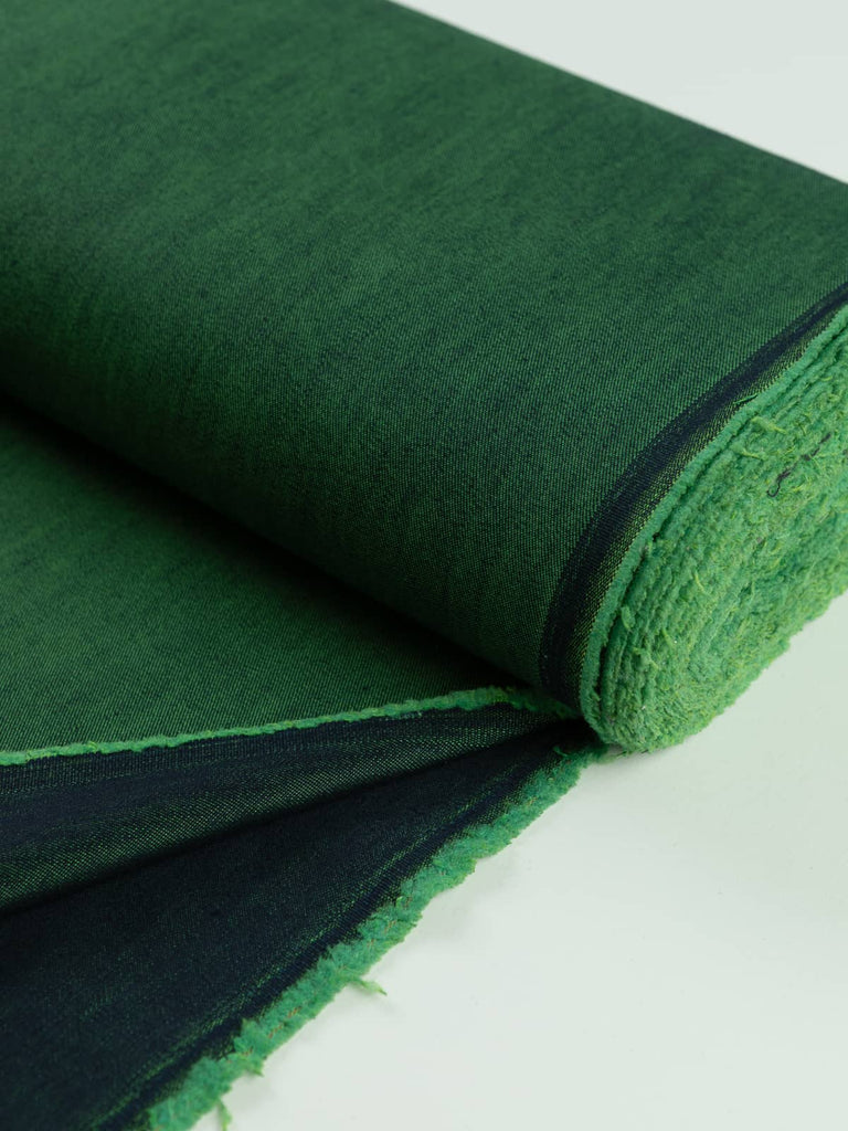 A neatly rolled piece of Emerald Gleam Denim fabric, highlighting the contrast between its rich navy warp and shimmering emerald weft. The fabric’s structured yet soft cotton-polyester blend is evident, with a lustrous finish catching the light.