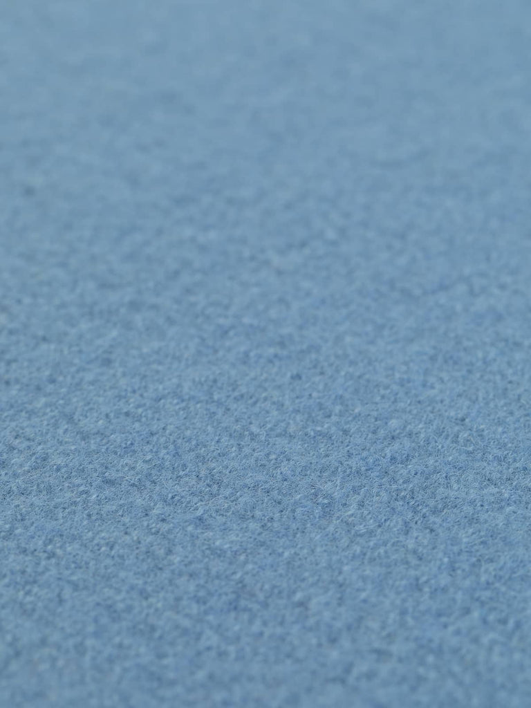 Close-up angled shot of Iona Sky boiled wool fabric laid flat, highlighting its gentle blue color and soft texture. Perfect for cozy garments with a hint of dreamy blue—order this versatile fabric today.